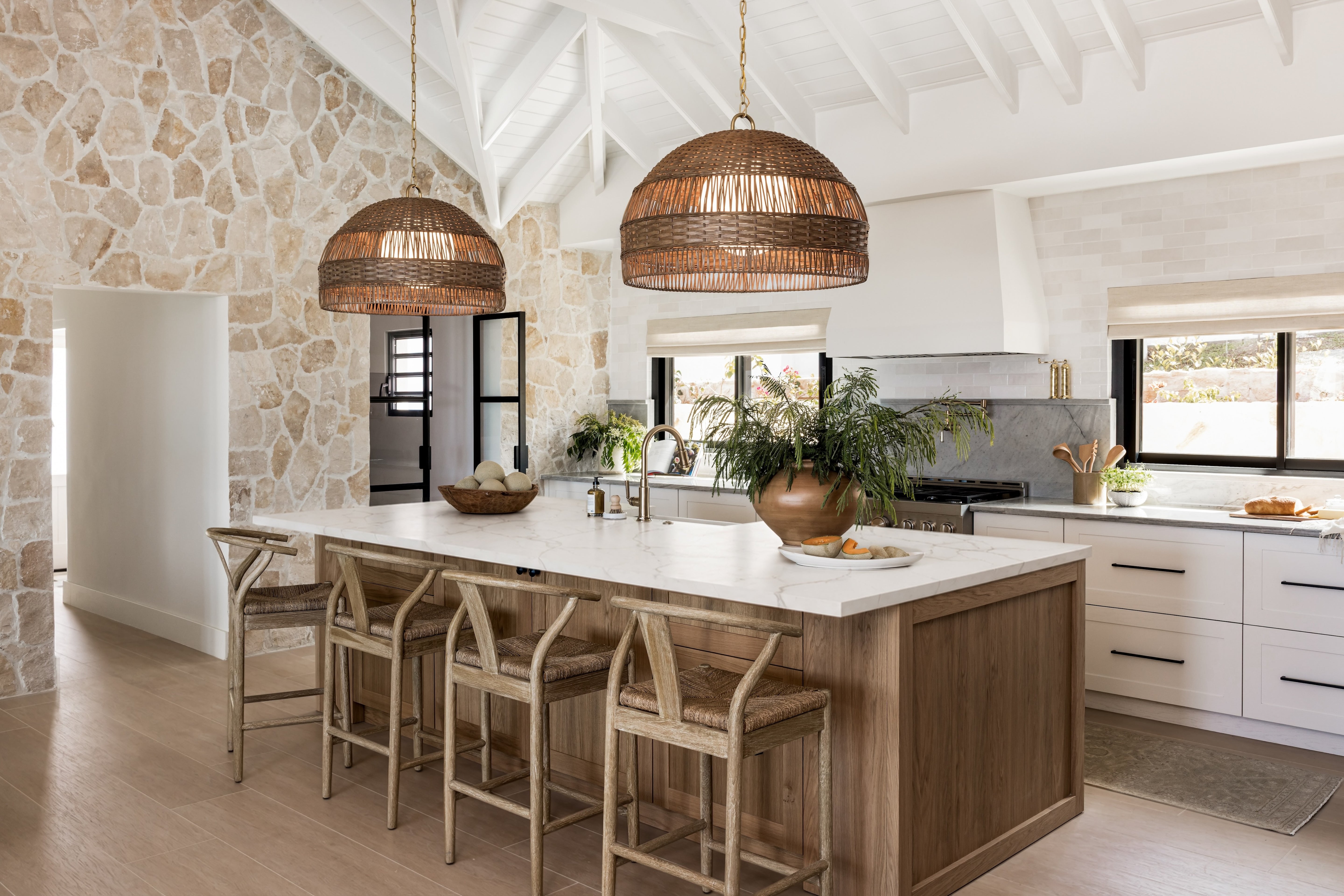 A modern kitchen island with a white countertop and wood base, surrounded by four light wood bar stools. Two large woven wicker pendant lights hang above the island.