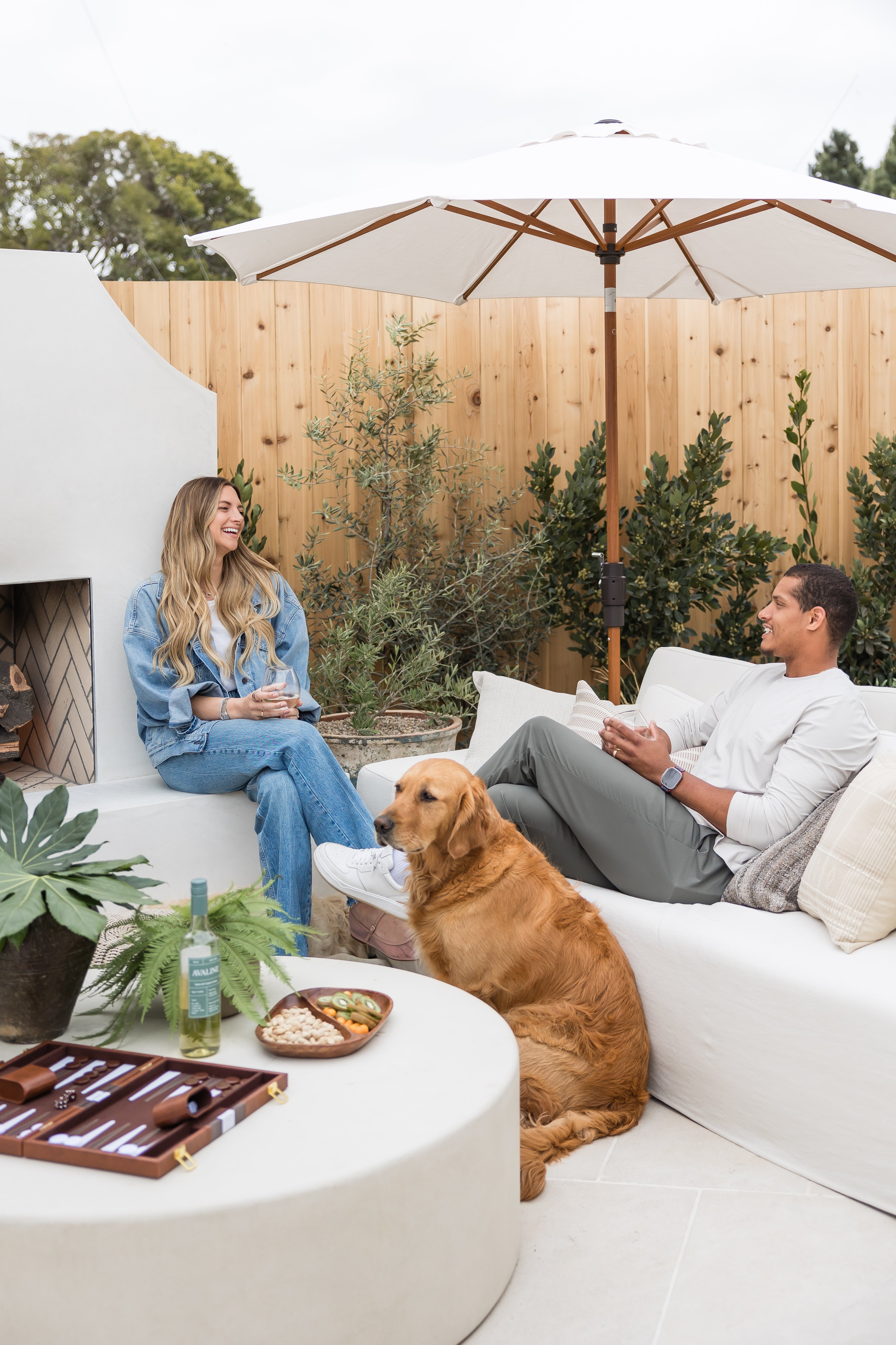 Two people sitting on a white couch with a dog, enjoying a casual outdoor setting.